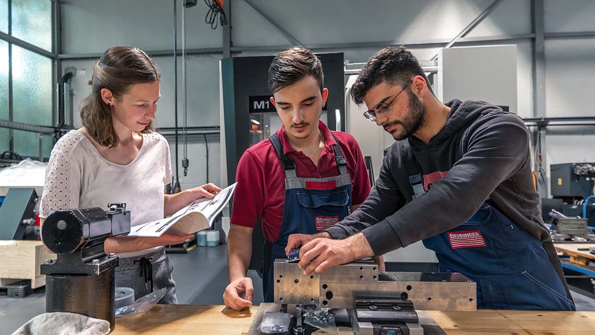 Eine Auszubildende zur Industriekauffrau und zwei technisch-gewerbliche Auszubildende stehen an einer Werkbank.